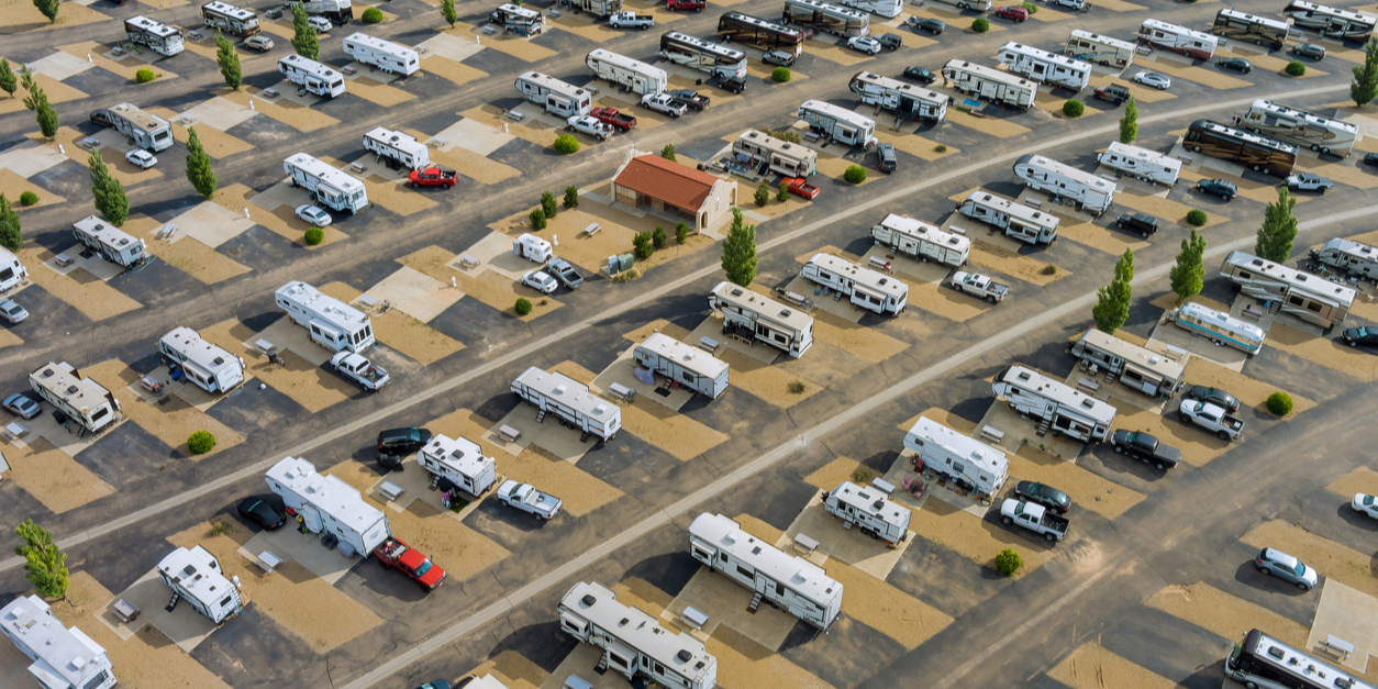Aerial view of an RV park with rows of parked RVs and cars, a small building in the center, surrounded by paved and sand areas, trees dotting the landscape.
